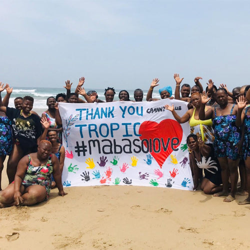 Image shows a group of women on the sand by the beach smiling holding a banner that reads: Thank you Tropic with a heart and handprints ~mabasolove